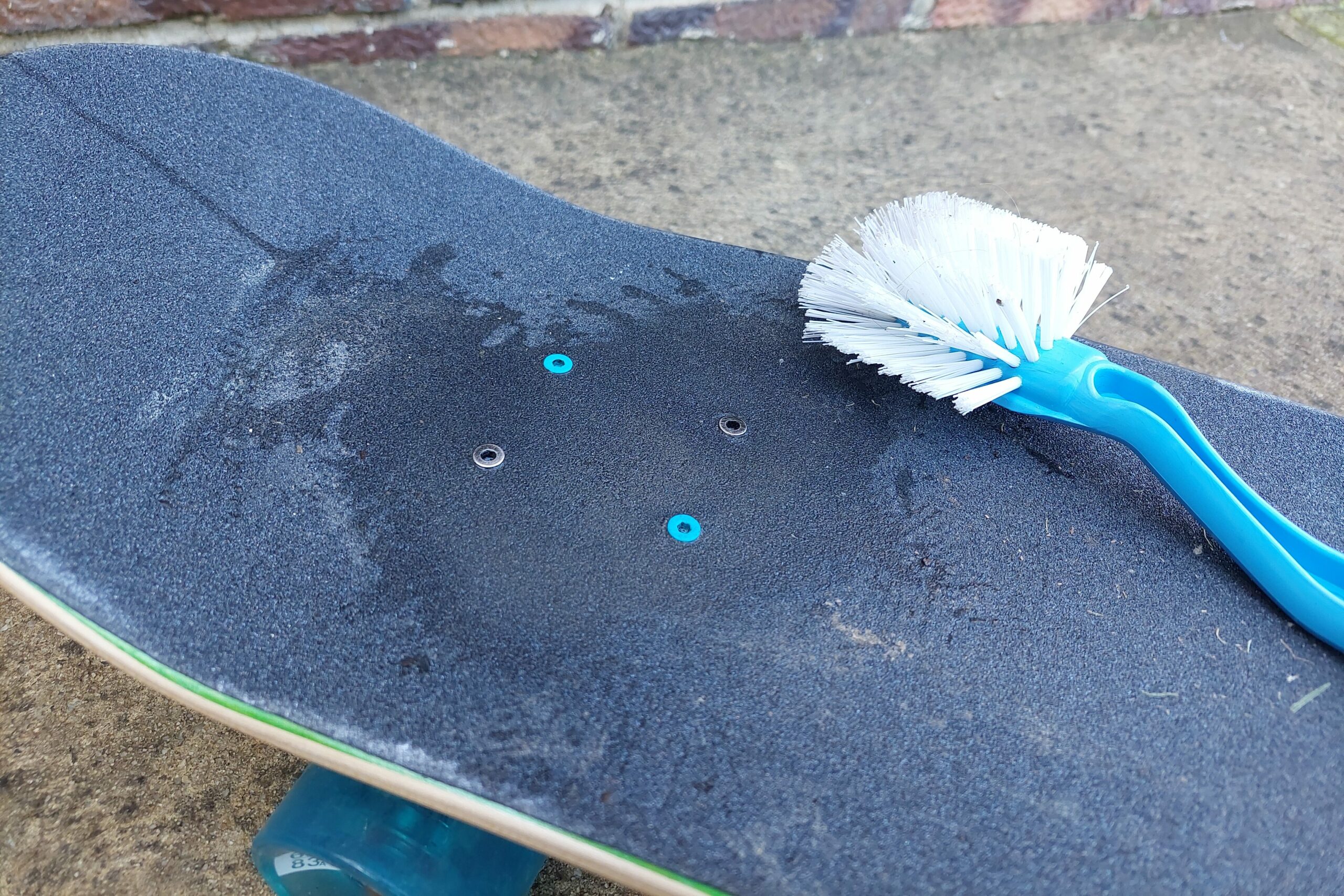 A close up of a dirty skateboard. There is a wet patch on the grip tape over the bolts where some of the dirt has been scrubbed off. The blue dishwashing brush is sitting on top of the board.