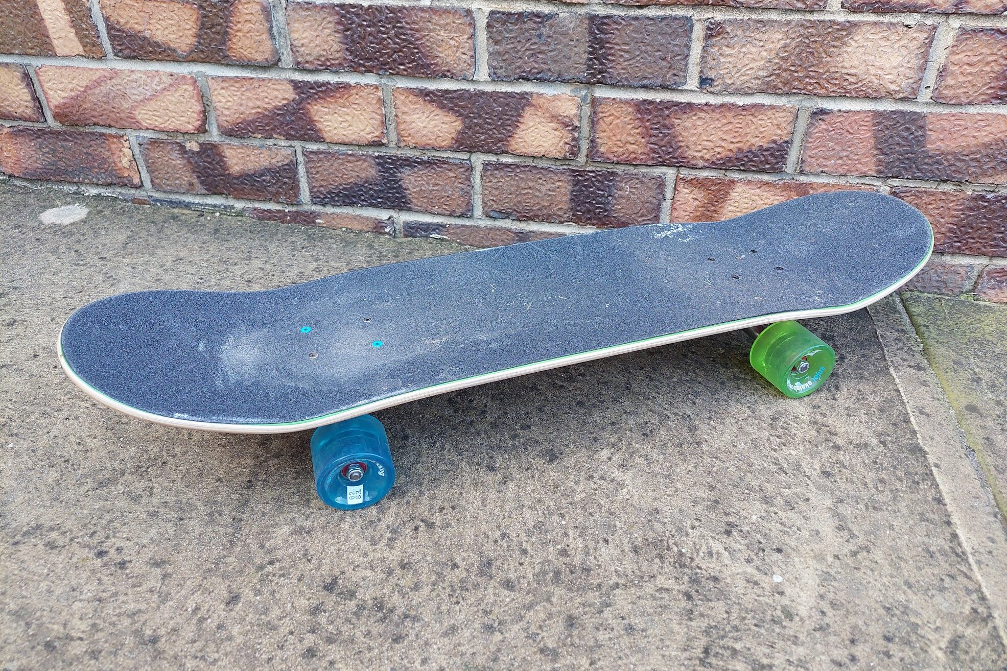 A dirty skateboard with black grip tape and blue and green wheels is sitting stationary on concrete with a brick wall in the background.